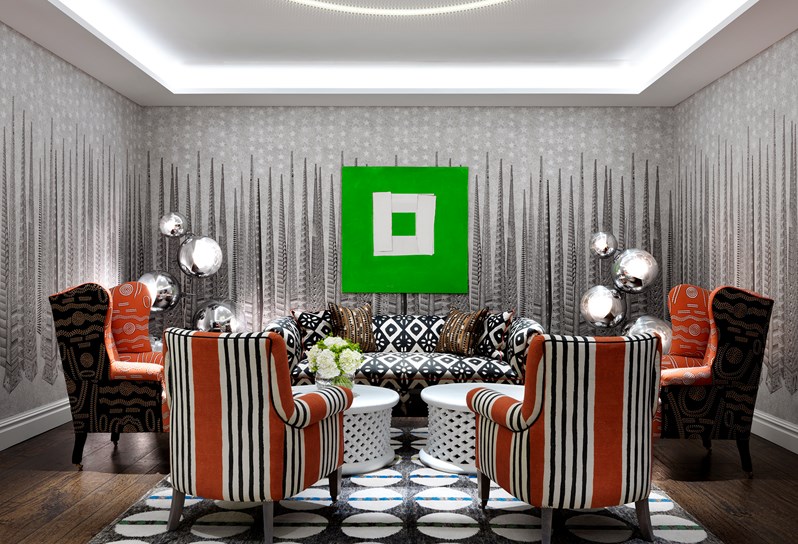 The Sandra Blow room at Soho Hotel. A grey graphic wallpaper and a black and white graphic carpet on the floor. There is a set of orange striped chairs, a black and white sofa and orange chairs are set around a coffee table. On the back wall is a bright green painting by Sandra Blow.