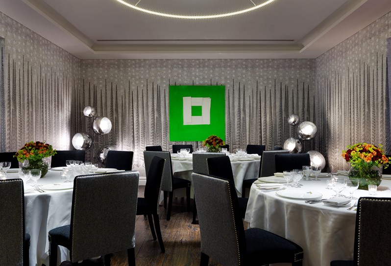 The Sandra Blow room at Soho Hotel. A grey graphic wallpaper and a black with oak floor. There is a is a bright green painting by Sandra Blow on the wall and three round tables set up for a private dinner with white linens on it.