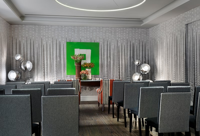 The Sandra Blow room at Soho Hotel. A grey graphic wallpaper and a black and an oak floor. It is set up theater style with grey backed chairs. There are two orange striped chairs at the front of the room with a table with flowers behind it . On the back wall is a bright green painting by Sandra Blow.