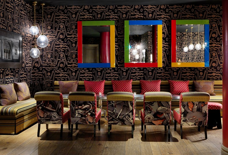 The Crimson Bar at Soho Hotel with a dark patterned wallpaper with multicoloured mirrors set upon the back wall. Banquette seating with tables run along the back wall.