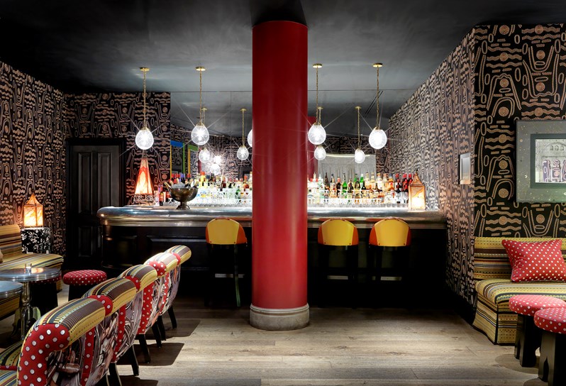The Crimson Bar at Soho Hotel. Dark patterned wallpaper on the walls, a bright red column runs down the centre of the room. In the background is a pewter bar with red and yellow bar stalls.