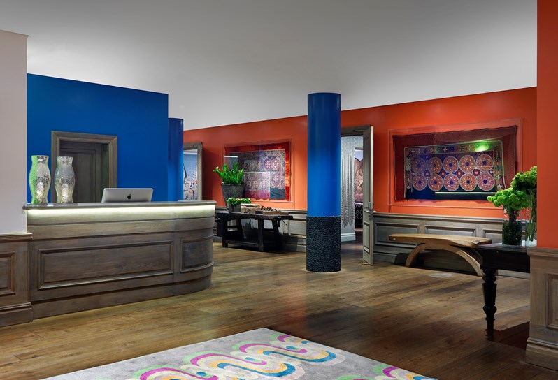 The reception area of the event floor for The Soho Hotel. It is open plan with oak wood floors with a colorful rug. There is a large reception check in desk to the left with two hurricane lamps on it. The walls of the space are in a cobalt blue and burnt orange. Framed tapestries hang on the wall