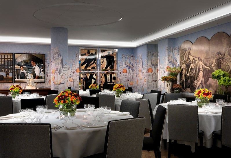 The Indigo Room, featuring light blue walls with an artistic pattern.  Round tables draped in white linen, formal china and colorful floral centerpiece fill the room beautifully. Candlelit votives and additional florals spread throughout the space. A five tier wedding cake on display set for an evening wedding reception dinner.