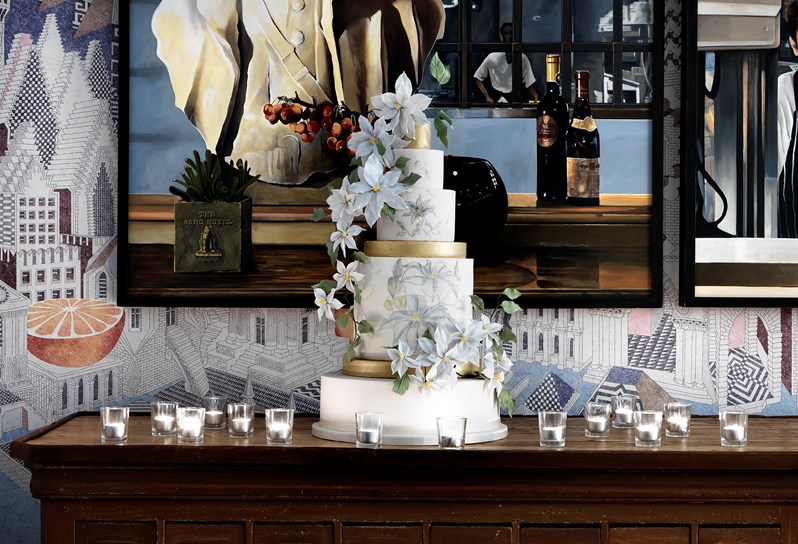 A five tier wedding cake displayed on a wood console set against the artistic pattern walls. Candlelit votives surrounding the cake to compliment the room setup.