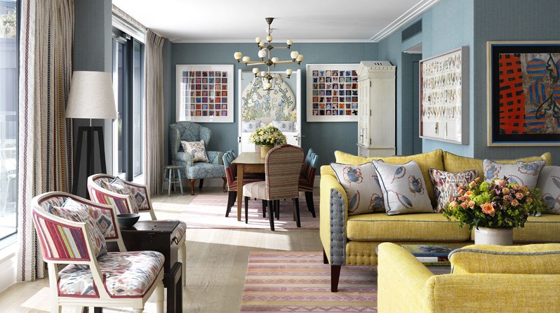 Ham Yard Hotel's Terrace Suite. A spacious seating area looking in toward a colorful bedroom, with colorful artwork on each side of the doorway, featuring lots of comfortable seating in two colorful armchairs and heavily patterned sofa places around a wooden coffee table, all with the light from floor to ceiling windows on the left.