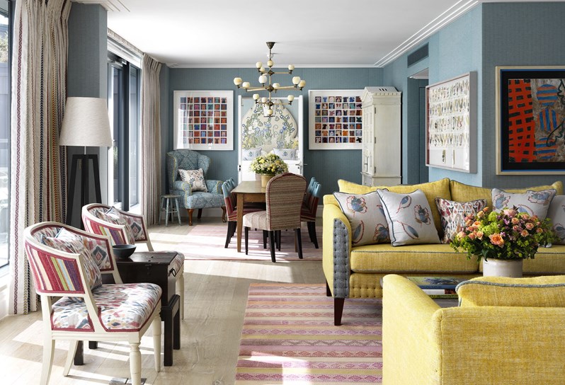 Ham Yard Hotel's Terrace Suite. A spacious seating area looking in toward a colorful bedroom, with colorful artwork on each side of the doorway, featuring lots of comfortable seating in two colorful armchairs and heavily patterned sofa places around a wooden coffee table, all with the light from floor to ceiling windows on the left.
