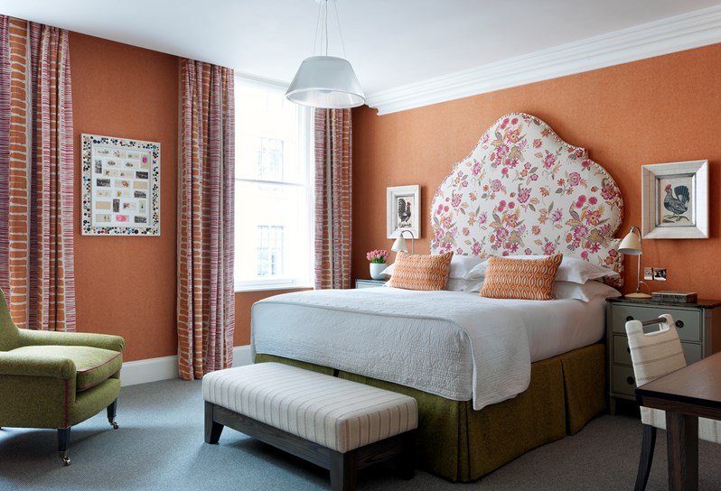 A bedroom in warm shades of orange, pink and white with a bold floral headboard and colorful curtains. There is a green arm chair and art featuring roosters.