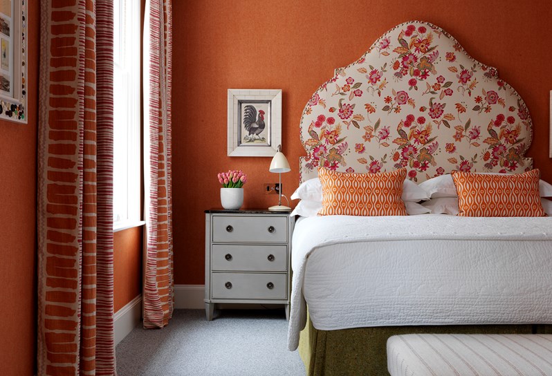 A close up of a bedroom in warm shades of orange, pink and white with a bold floral headboard and colorful curtains. The bedside table has flowers and a reading lamp and there is art featuring roosters.