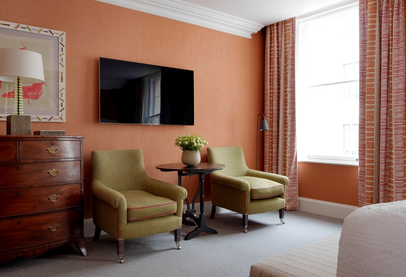Sitting area of the bedroom with two green arm chairs. There is an antique dresser against the burnt orange walls and colorful curtains in shades of orange, white and pink.
