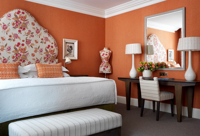 Bright bedroom in warm shades of orange, pink and white with a bold floral headboard and mannequin. The writing desk has colorful flowers on it and two tall lamps as well as a mirror.