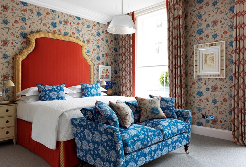A boldly colored bedroom with floral patterned walls in shades of tan, red and blue. There is a red headboard with yellow trim and bright blue and white patterned throw pillows and couch.