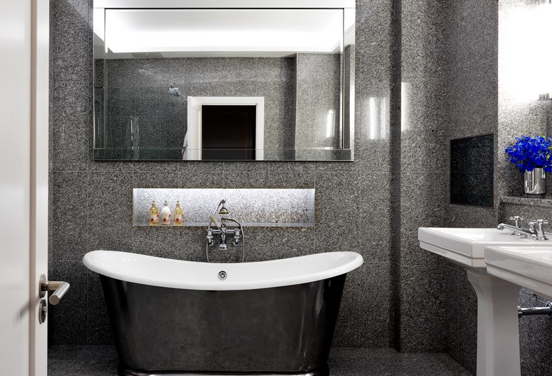 A beautiful granite bathroom with a deep soaking tub, large mirror above the tub and a television. To the right are two basins.