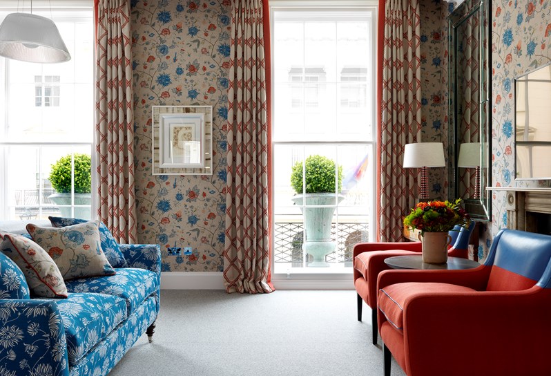 A view facing the windows in a boldly colored bedroom with floral patterned walls in shades of tan, red and blue. The two windows are floor to ceiling with large topiaries outside. To the left is a beautifully printed blue and white couch and to the right in front of the fireplace are two chairs in red and blue.