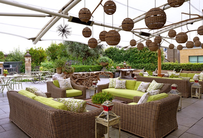 A semi covered, leafy roof terrace; two seating arrangements with rattan sofas and armchairs with  fluffy lime green cushions and coffee tables under a canopy with quirky lanterns and further back an uncovered fountain and leafy garden.