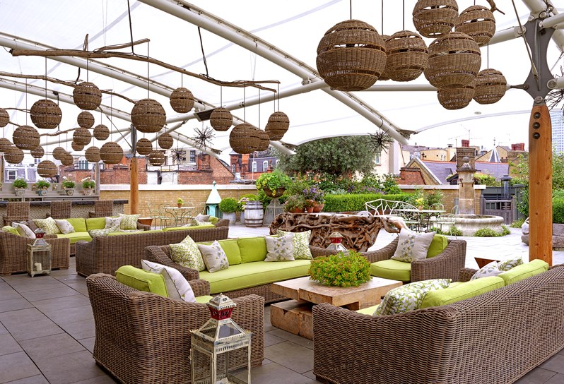 Two spacious seating areas featuring rattan armchairs and sofas with lime cushions and details arranged in front of a bar, all on a roof terrace partially covered with a white canopy with lots of quirky lanterns.