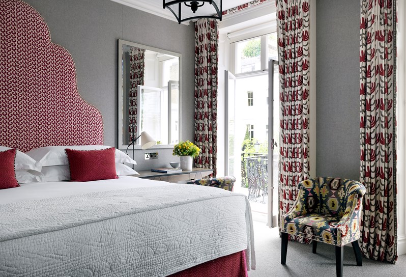 A grey walled large bedroom with a red headboard and large French doors which are open facing onto the street below.  There is desk next to the bed and a bucket style chair between the French doors in a multi coloured patterned fabric.