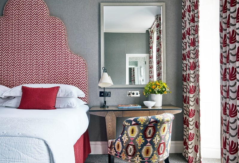 A close up image of a double bed with a red patterned headboard against a grey wall.  To the right there is a wooden desk with a bucket style chair in a patterned fabric.  There are curtains to match the viewed to the side of a window.