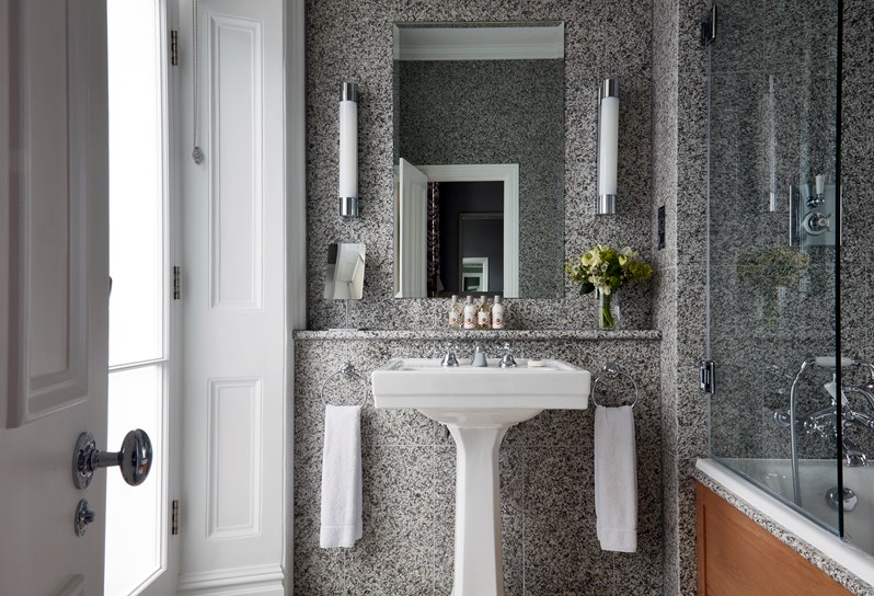 A bathroom in grey granite with a pedestal basin in white ceramic and large mirror above the sink. There is a bath and overhead shower to the right.  A window to the left.
