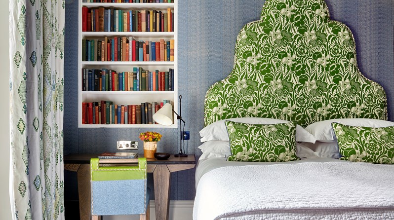 A bedroom in a light blue colour showing a bed straight on with a green patterned headboard. To the left of the bed there is a wall handing book shelf with a wooden desk underneath with a green and blue chair.