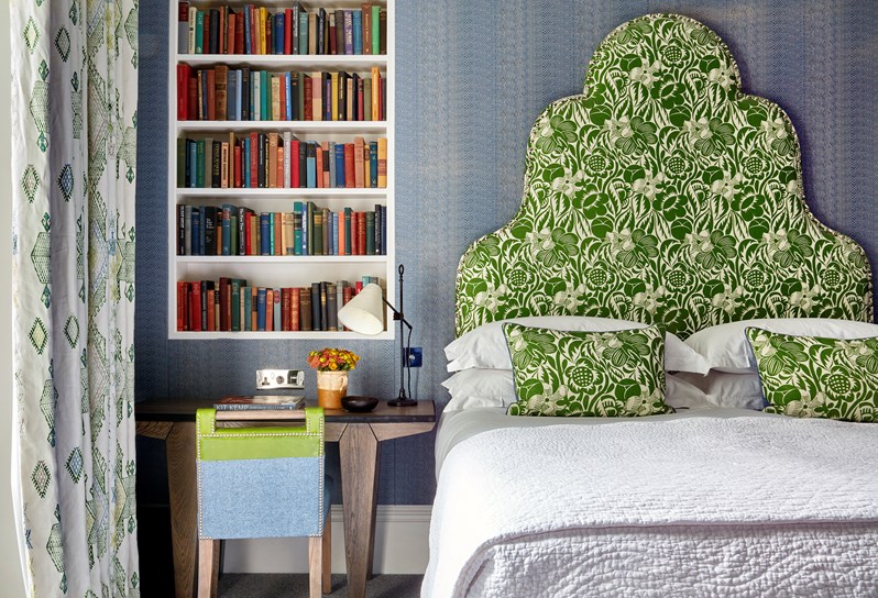 A bedroom in a light blue colour showing a bed straight on with a green patterned headboard. To the left of the bed there is a wall handing book shelf with a wooden desk underneath with a green and blue chair.