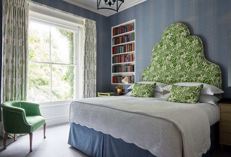 A spacious bedroom with a large double bed with a patterned green headboard.  There is a window and book shelf and a bucket seat in green just under the window.