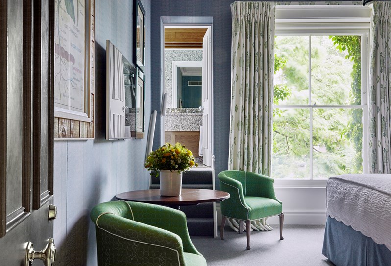A view from the entrance of the bedroom facing the large window.  There is a wooden round table and two green bucket style chairs against the wall.  A small staircase leads up to the bathroom.