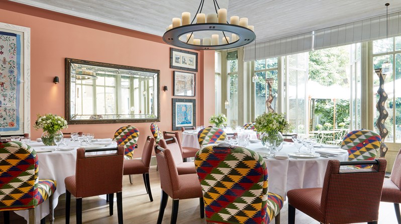 The sunny Orangery at Number 16 with salmon pink walls and wooden floorboards.  Three round tables in the centre of the room are covered in white table cloths and laid for a dinner service.