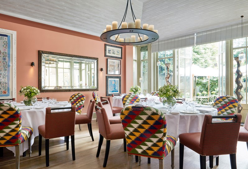 The sunny Orangery at Number 16 with salmon pink walls and wooden floorboards.  Three round tables in the centre of the room are covered in white table cloths and laid for a dinner service.