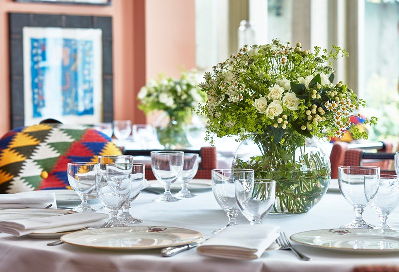 Close up image of a table set for dinner with glasses and Kit Kemp's mythical creatures plates.  There is a large flower arrangement in the centre of the table.