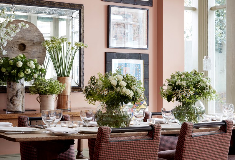 A close up of a set table in the orangery with beautiful flowers in green and white on the table and vases with further flowers on the side table.  The room featured hand picked art.