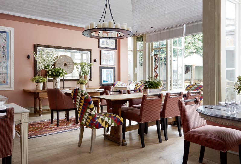 The image of the orangery at Number Sixteen showing various dining tables with brightly coloured chairs and wooden and marble tables. There are vases with white and green flowers and individual hand picked art on the walls.  The room faces out onto the garden with large windows.