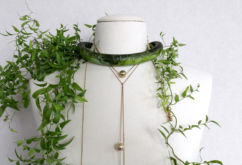 A white mannequin with leafy plants around and on the shoulders, wearing a moss neck ring and long gold necklaces.