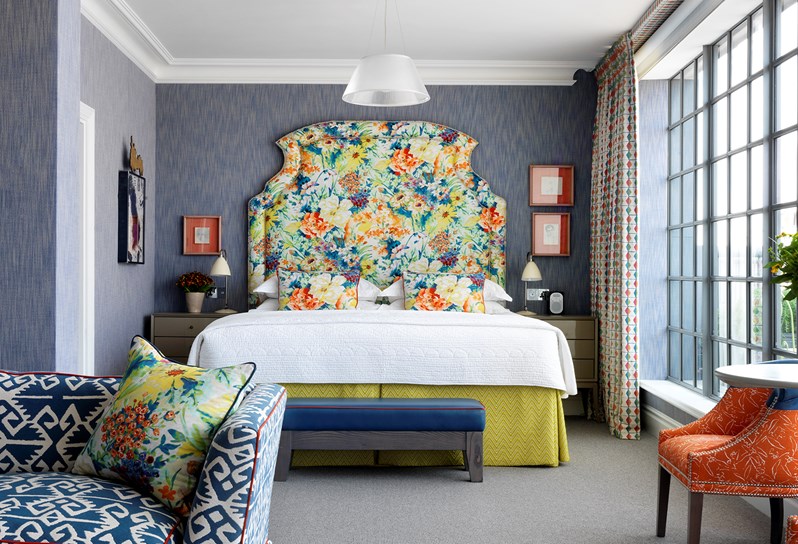 A stunning room with blue walls and a brightly patterned floral headboard.  There are orange chairs in front of the floor to ceiling windows and a blue and white couch in the forefront.
