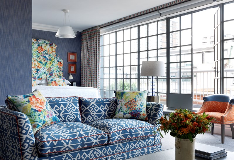 A stunning room with blue walls and a brightly patterned floral headboard.  There are orange chairs in front of the floor to ceiling windows and a blue and white couch in the forefront with the same floral pattern on the throw pillows.