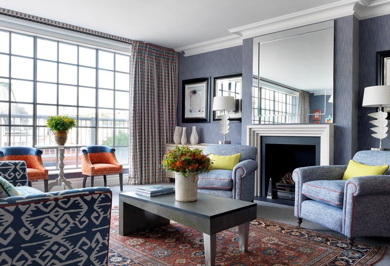 A view of the living room in a stunning room with blue walls. There are orange chairs in front of the floor to ceiling windows and a blue and white couch faces the fireplace. Underneath the coffee table is a beautiful Persian rug