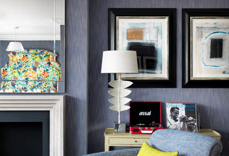 A close up view of details in the living room. On a table next to the fireplace is a record player and decorative lamp with two pieces of watercolor art behind.