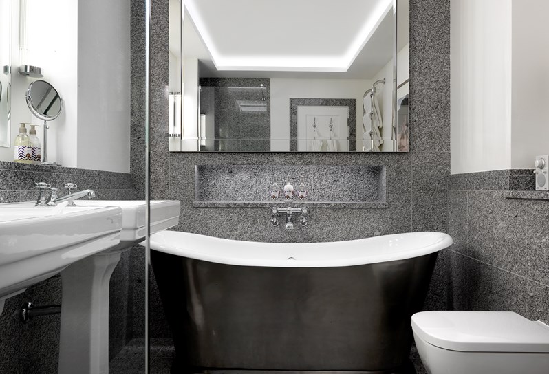 A bathroom with grey granite walls. A walk in shower and in the middle of the room a cast iron soaking tub. Above the tub is a larger mirror. There are two white basins in the bathroom