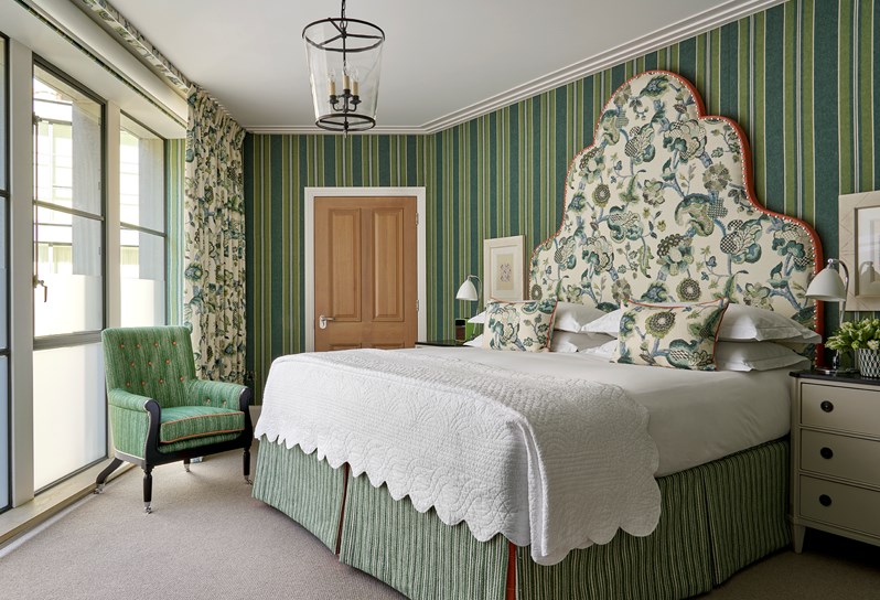 An image of the bedroom of The Deluxe Two Bedroom Suite, Room 5 . A stylish bedroom featuring a vibrant patterned headboard and pillows, green textured wallpaper, eclectic artwork and floral arrangement.