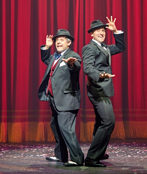 Two men dressed in suits and fedoras stand back to back singing on stage in The Producers