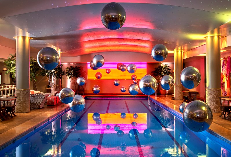 A lot of large Metallic looking baubles hanging from the ceiling over the pool with a projected background of colours on the wall behind. There are two poolside tables visible on the right hand side, and a sofa area and poolside table on the left.