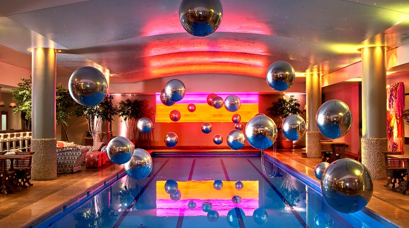 A lot of large Metallic looking baubles hanging from the ceiling over the pool with a projected background of colours on the wall behind. There are two poolside tables visible on the right hand side, and a sofa area and poolside table on the left.