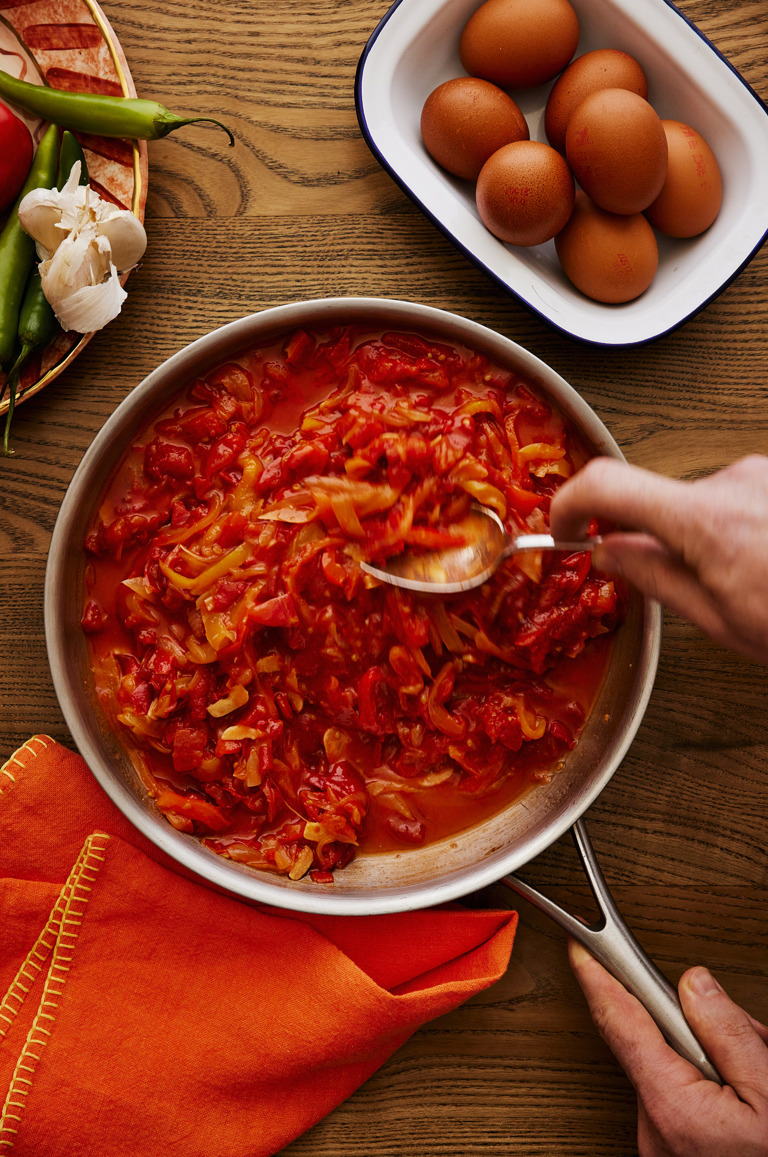 This is a portrait image showing the stage of making the tomato based sauce in which the eggs will be baked. It shows a silver frying pan with someone stirring the tomato based sauce with a spoon.  in the back of the image you can see a small blue and white baking tray filled with eggs and to the left a Sasha Compton dish filled with fresh vegetables.  