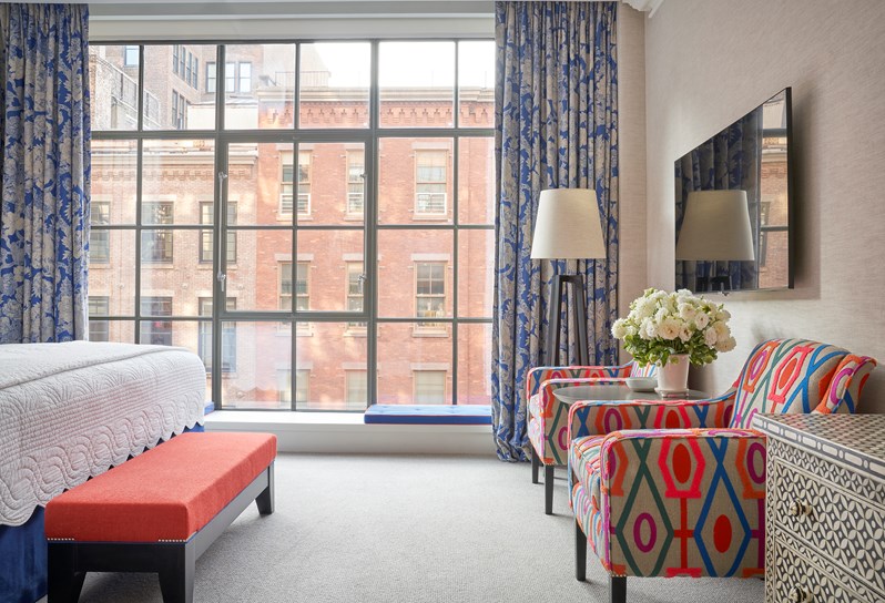 Shot of the bedroom in a Deluxe Room in The Crosby Street Hotel facing a large window with the end of the bed on the left of the picture and two colourful arm chairs on the right by a wall with a tv screen above.