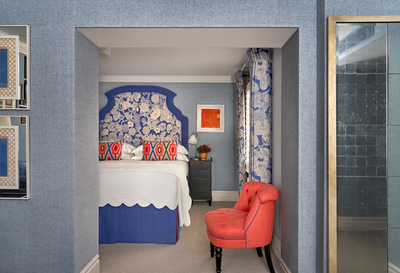 Bedroom shot of room 15, the Marylebone Room at Dorset Square Hotel. The image shows the bedroom area, on the left, the bed with a large blue and floral statement headboard . on the right matching curtains and a coral tub chair.