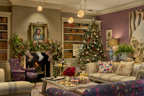 The library at Haymarket Hotel decorated for Christmas. There are canapes, Champagne and presents on a coffee table between the sofas. There is a garland and stockings over the fireplace with a bar cart holding glasses and bottles in front. In the corner there is a decorated Christmas tree