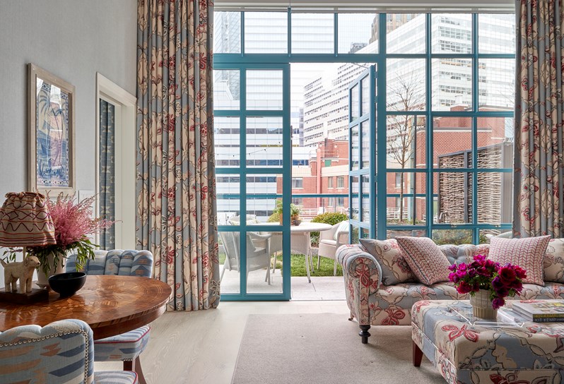 A living room area. A footstall in blue 'Chainstitch' fabric which matches the sofa opposite and curtains on the floor to ceiling windows and doors that open up onto a landscaped terrace with woven seating and a table. Inside on the left, two blue patterned chairs with a wooden table in the middle with a doggy lamp and flowers on.