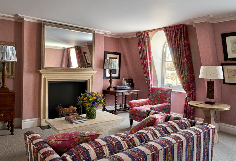 A lounge area with a fora and armchair facing a coffee table in the center and a flower on it with a big mirror and a fire place. A big windows streaming light in to the side.