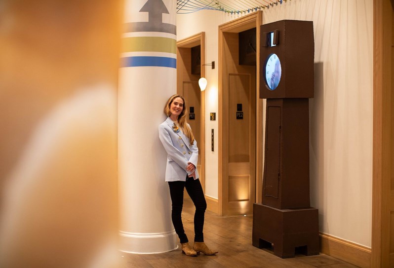 Firmdale Hotels Art Ambassador and Design Director, Willow Kemp stands leaning against a column in the lobby at The Whitby Hotel. To her left, a piece of artwork by Humans Since 1982 stands against the wall.