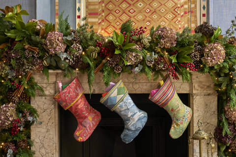 This image shows a close up of a festive fireplace in the Townhouse at Haymarket Hotel, decorated with a Christmas garland featuring hydrangeas, red berries, pinecones, cinnamon sticks, seasonal greenery and twinkling lights, with three colourful patterned stockings hanging from the mantelpiece.