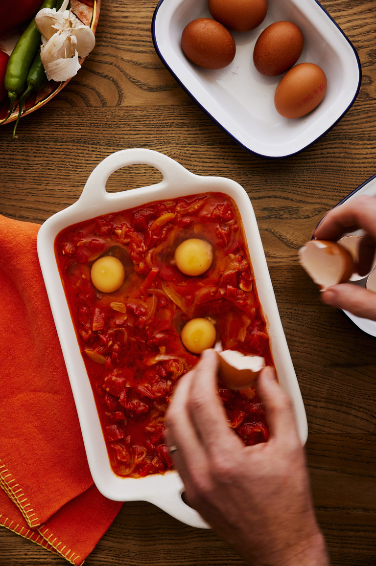 In this image you can see hands dropping eggs into wells made in he tomato sauce preparing to bake, there is a white baking dish filled with red tomato based sauce and a bowl of eggs can be seen in the top of the image. 